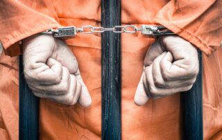 handcuffed-hands-of-a-prisoner-behind-the-bars-of-a-prison-with-orange-clothes-crispy-desaturated